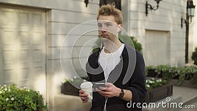 Giovane con le cuffie ed il caffè che cammina nella via video d archivio Giovane con le cuffie ed il caffè che cammina nella via video d archivio