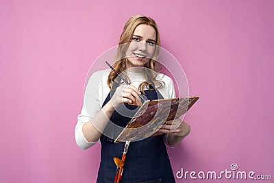 Girl artist holds brushes and a palette and smiles on a pink background, student of art school, profession of an artist Stock Photo