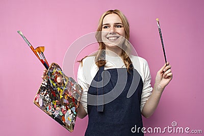 Girl artist holds brushes and a palette and smiles on a pink background, student of art school, profession of an artist Stock Photo
