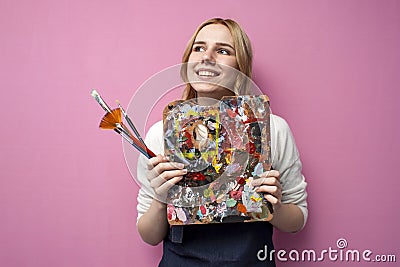 Girl artist holds brushes and a palette and smiles on a pink background, student of art school, profession of an artist Stock Photo