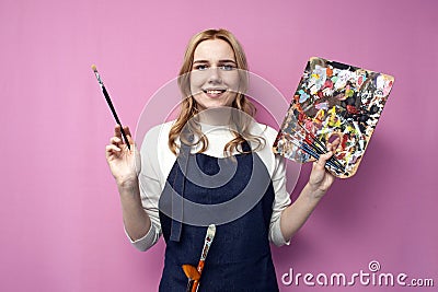 Girl artist holds brushes and a palette and smiles on a pink background, student of art school, profession of an artist Stock Photo