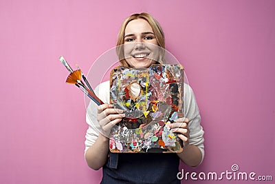 Girl artist holds brushes and a palette and smiles on a pink background, student of art school, profession of an artist Stock Photo