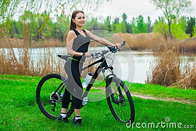 Girl athlete with blond, long hair in black sports tights and T-shirt walking in the morning with the bike in the park Stock Photo