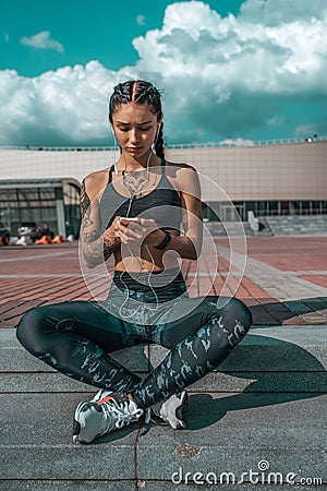 Girl athlete, summer city. Listens to music on headphones, chatting in social networks. Sits in leggings sneakers. Woman Stock Photo