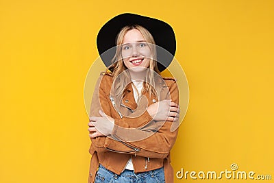Girl in autumn warm clothes hugs herself with hands and smiles on a yellow isolated background, dress warmly concept Stock Photo