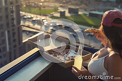 A girl with a beautiful breast in a sports suit eats pizza and drinks mojito on the sunset city background Stock Photo