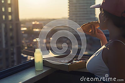 A girl with a beautiful breast in a sports suit eats pizza and drink mojito on the sunset city background Stock Photo