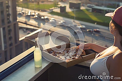 A girl with a beautiful breast in a sports suit is going to eat pizza and drink mojito on the sunset city background Stock Photo