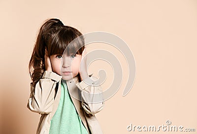 A girl in a beige coat stands with her hands over her ears. Stock Photo