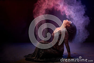 Girl in black dress posing in dark studo during photoshoot with flour or dust and light. Dangerous witch during struggle between Stock Photo