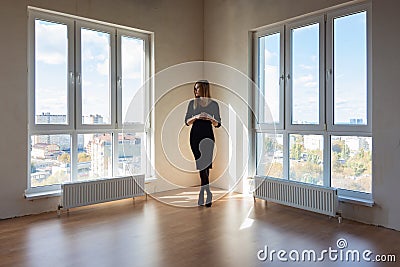 A girl in a black dress stands between two large windows in a spacious empty apartment Stock Photo