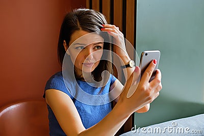 A girl in a blue dress sits at a cafe uses the front camera of the phone as a mirror Stock Photo