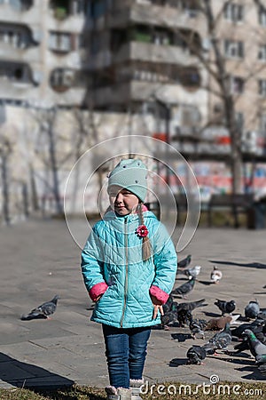 A girl in a blue jacket poses outside in front of a flock of city pigeons. A five-year-old child with long pigtails. Blurred image Stock Photo