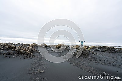 Girl with blue jacket are standing on a sand dune with arms raised in Stokksnes and celebrates the view. Stock Photo