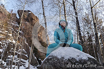 Girl in blue sportswear sitting on a large boulder on the nature on the background of rocks in the winter Stock Photo