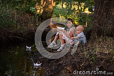 Girl and boy are sitting on the bank of river, launching white paper origami boat into the water Stock Photo