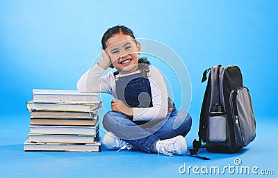 Girl child, sitting and books in studio portrait, backpack and excited for learning on floor by blue background. Female Stock Photo