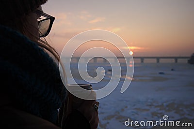 A girl with coffee paper cup in her hands is enjoying sunrise on bank of River Dnipro. Close-up photo. Stock Photo