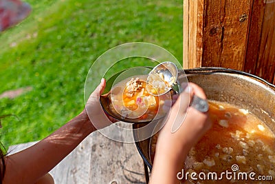 The girl cook outdoors puts soup with meat and vegetables borscht from a large cauldron pan on the wooden floor Stock Photo