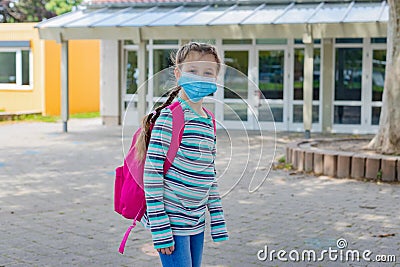 A girl with a crimson, pink backpack stands in front of the school doors in a protective mask. Back to school, starting school Stock Photo