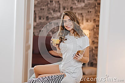 Girl with cute smile enjoying orange juice while listening music on couch. Indoor portrait of glad beautiful lady in Stock Photo
