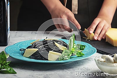 Girl cuts cheese to decorate black spaghetti dishes. Unusual cuttlefish ink pasta. A wholesome light dinner with basil Stock Photo
