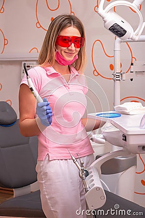 A girl dentist wearing red glasses and a photo lamp in her hands, smiling in the dental office. Stock Photo