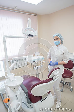 Girl dentist, in white clothes, mask and cap stands next to a modern dental chair in a large, simple, medical office Stock Photo