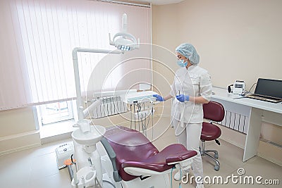 Girl dentist, in white clothes, mask and cap stands next to a modern dental chair in a large, simple, medical office Stock Photo