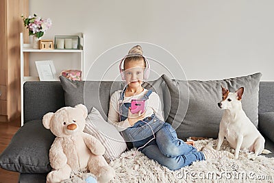 Girl and dog laying on couch in headphones, listening to music with her smarthphone. Little girl watching cartoons on her phone. Stock Photo