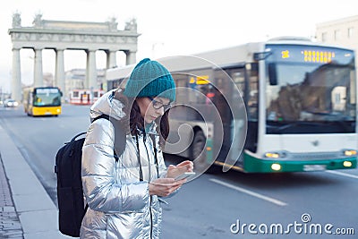 Girl in a down jacket and hat stands at a public transport stop and uses a smartphone. Wait for the bus. Stock Photo