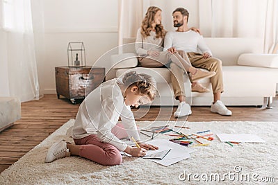 Girl drawing and sitting on floor, parents sitting on sofa behind Stock Photo