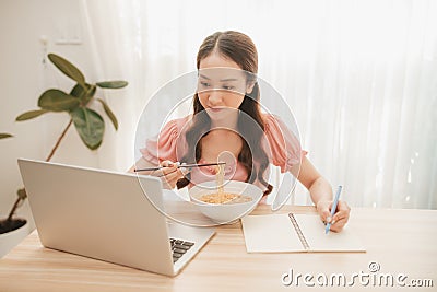 Girl eating instant noodle in hurry time while running her business at home on notebook computer Stock Photo