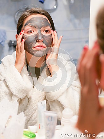 The girl in the face mask looks in the mirror displeased. Cosmetic black mask. Woman brings beauty to her face, gets rid of black Stock Photo