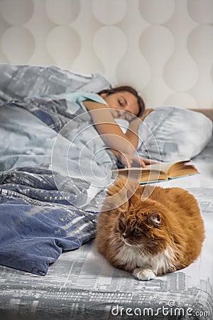 The girl fell asleep in a cozy bed reading a book. Along with the woman on the bed lies a fat, fluffy red cat Stock Photo