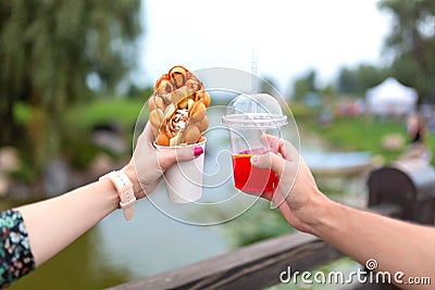 The girl and fella during the walk holds in hands a paper cup with a Belgian waffle and fresh drink on a background of green park Stock Photo