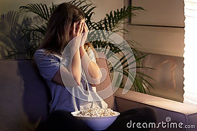 Girl with a frightening face is watching TV and covered her face with her hands. Stock Photo