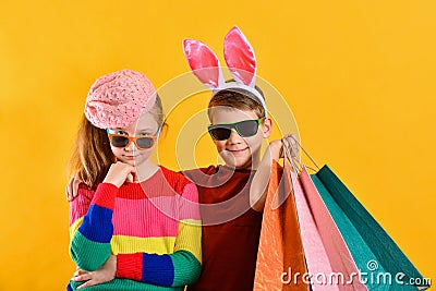 A girl with glasses and a boy with rabbit ears are bought for Easter. Children with colored bags for goods on a yellow background Stock Photo