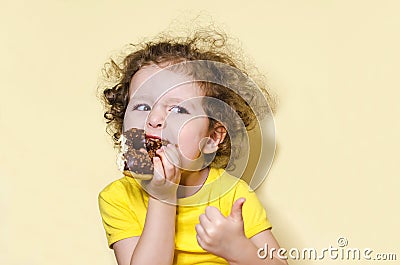 Girl with greed and appetite eats, bites delicious chocolate donut and raises finger up on yellow background Stock Photo