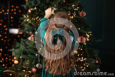 Girl in green dress and with a bowknot of red ribbon on her hair is standing in living room from behind near Christmas tree. Stock Photo