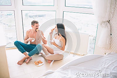 Girl and guy are sitting on floor with their legs crossed. They eat croissans and drink from white cups. Couple is Stock Photo