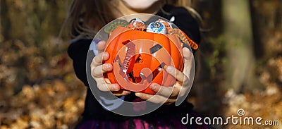 Girl in Halloween skeleton dress offers you orange pumpkin plastic bucket full of traditional candies and jelly worms. Stock Photo