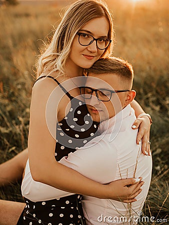 A girl in hat sits in hands guy on ground in middle of tall grass in meadow Stock Photo