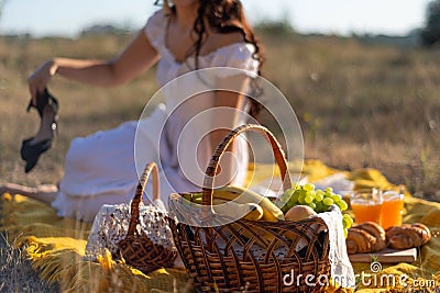 Girl in a hat and white dress sitting on a picnic in a grass field in defocus blur. On the cross plan are baskets with fruit. Stock Photo