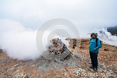 Girl with hiking gear infront of sulphur steam vents in Iceland during heavy cold wind. Mineral rich and textured ground infront. Stock Photo