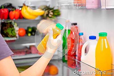 A girl holds a bottle of orange vegetable or fruit juice against the background of an open refrigerator with food. Concept of Stock Photo