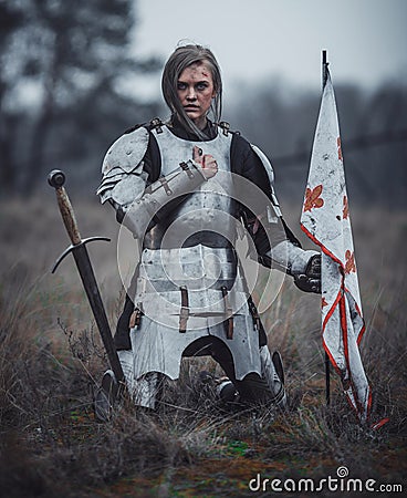 Girl in image of Jeanne d`Arc in armor kneels with flag in her hands and sword on meadow. Stock Photo