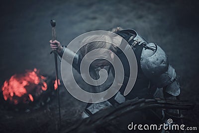 Girl in image of Jeanne d`Arc in armor and with sword in her hands kneels against background of fire and smoke. Stock Photo