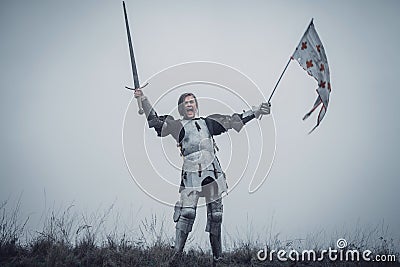 Girl in image of Jeanne d`Arc stands in armor and issues battle cry with sword raised up and flag in her hands. Stock Photo