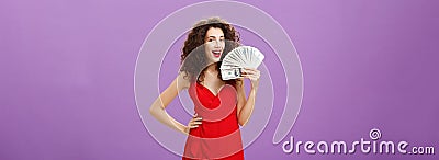Girl knows how earn money. Successful and delighted satisfied woman in elegant red dress with curly hair holding cash Stock Photo